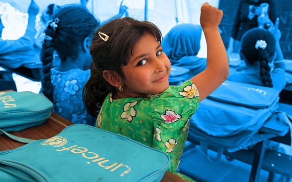 Una bambina si volta sorridendo mentre, insieme alle sue compagne, alza la mano per rispondere alla domanda della maestra.