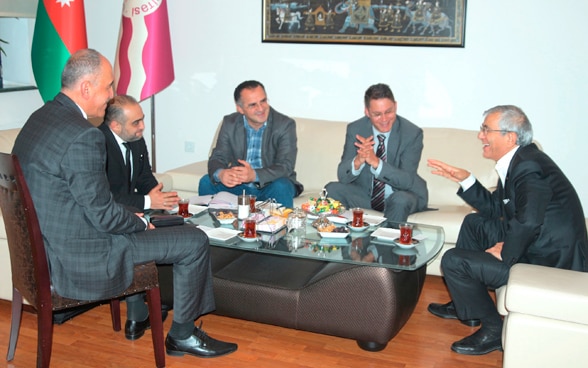 Five men having a discussion around a table in an official setting. 