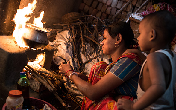 Eine indische Frau bereitet Essen auf einem Kochherd zu. Neben ihr steht ein Kind.