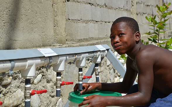 A young boy drinks water and refills a water bucket from taps installed by Swiss Humanitarian Aid.