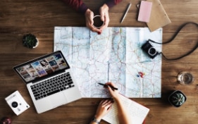 Two people at a table planning a trip with a map, stationery and a laptop. 