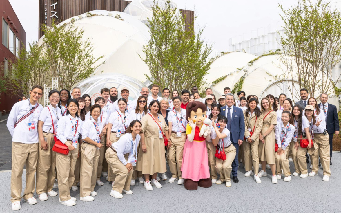 Un grand groupe de personnes en uniforme pose avec une personne déguisée en Heidi devant des bâtiments en forme de dôme et des arbres.