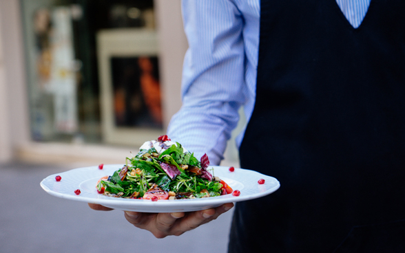 Un homme vêtu d’une chemise à rayures et d’un tablier sert une salade colorée.