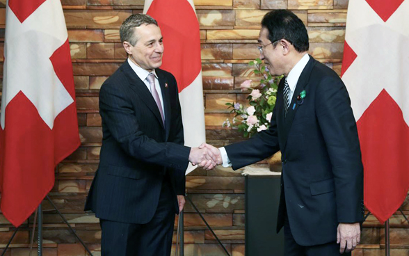 Fumio Kishida e Ignazio Cassis si stringono la mano. Sullo sfondo si vedono le bandiere del Giappone e della Svizzera. 
