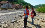 Two women in reflective vests looking at a destroyed house on the banks of a stream.