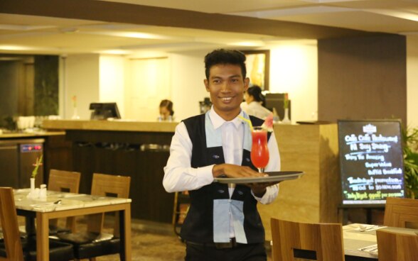 A young waiter smiles as he walks through a restaurant, holding a cocktail on a tray.