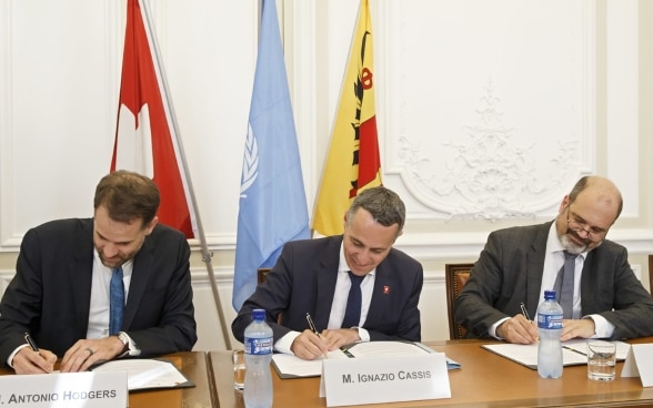 Le conseiller fédéral Cassis, Antonio Hodgers et Sami Kanaan s'assoient à une table en bois et signent la déclaration commune.