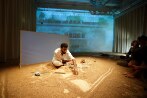 Un homme est assis sur la scène et dessine dans le sable, le public est assis très près de lui, à même le sol. Un film est projeté sur un écran derrière l’artiste.