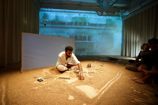 A man sitting on a stage drawing in sand, with the audience sitting on the floor very close by. A film is being shown on a screen behind the artist