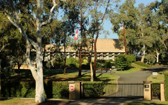 The Swiss embassy building set in a garden with a Swiss flagpole and a large tree in the foreground.