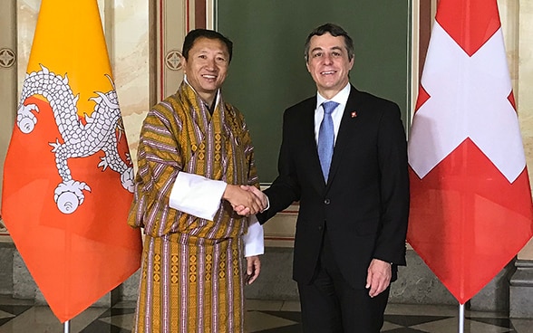 Ignazio Cassis shakes hands with the traditionally dressed foreign minister of Bhutan, Tandi Dorji. They are standing in front of the two states' flags.