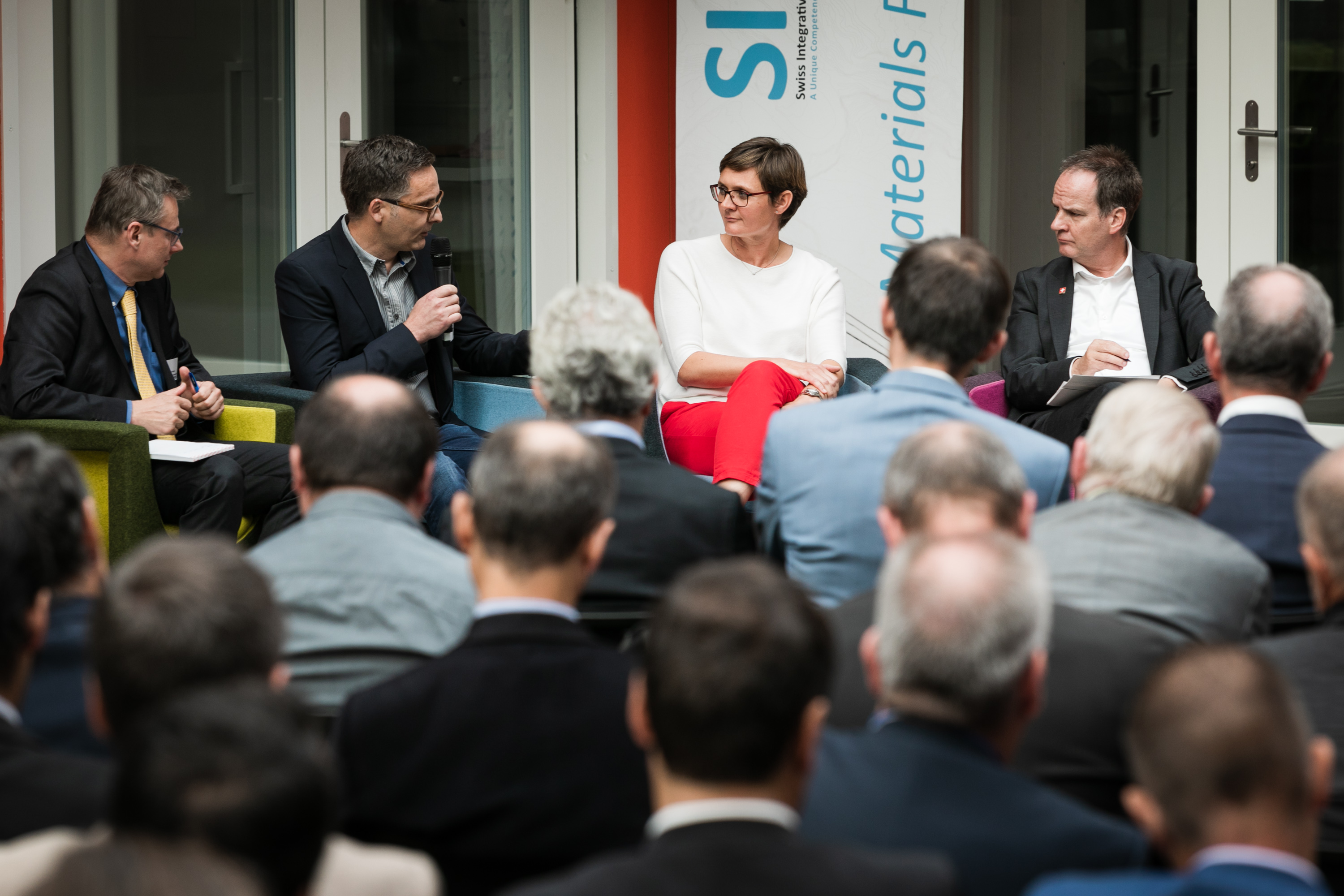 Discussion panel of men addresses questions to a young woman in red pants.