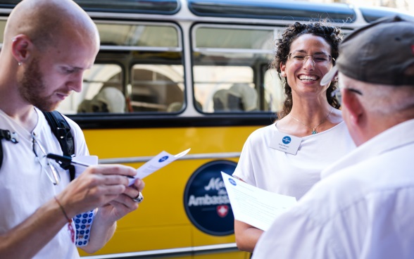 Une ambassadrice aux cheveux foncés parle à deux visiteurs; à l'arrière-plan se trouve le bus postal Meet the Ambassador