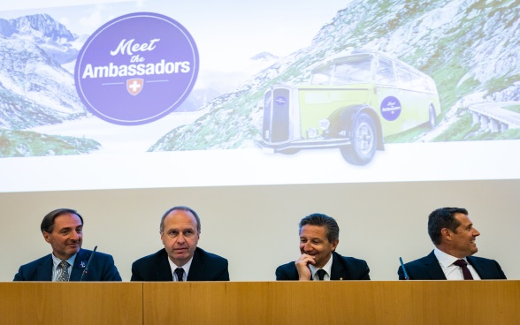Quatre ambassadeurs sur le podium de l'auditorium, à l'arrière-plan une photo du bus postal  Meet the Ambassador