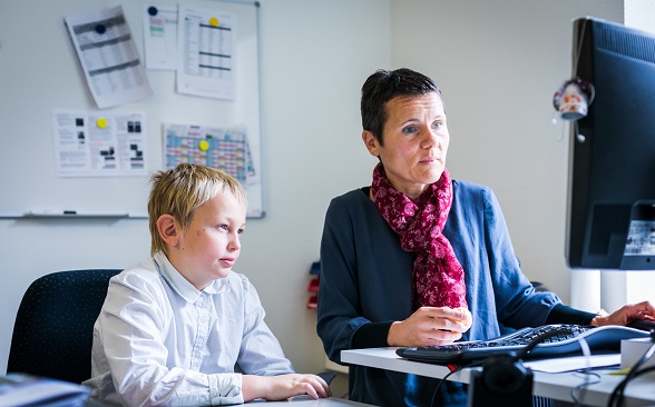 A boy watches a woman work at a computer.