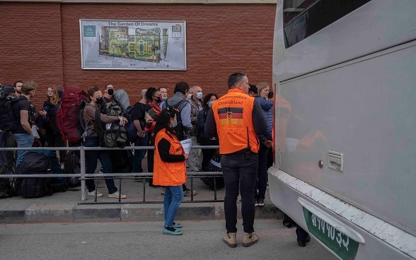 Tourists stand in line to board a bus. Two members of the German Embassy in Kathmandu are standing next to it.