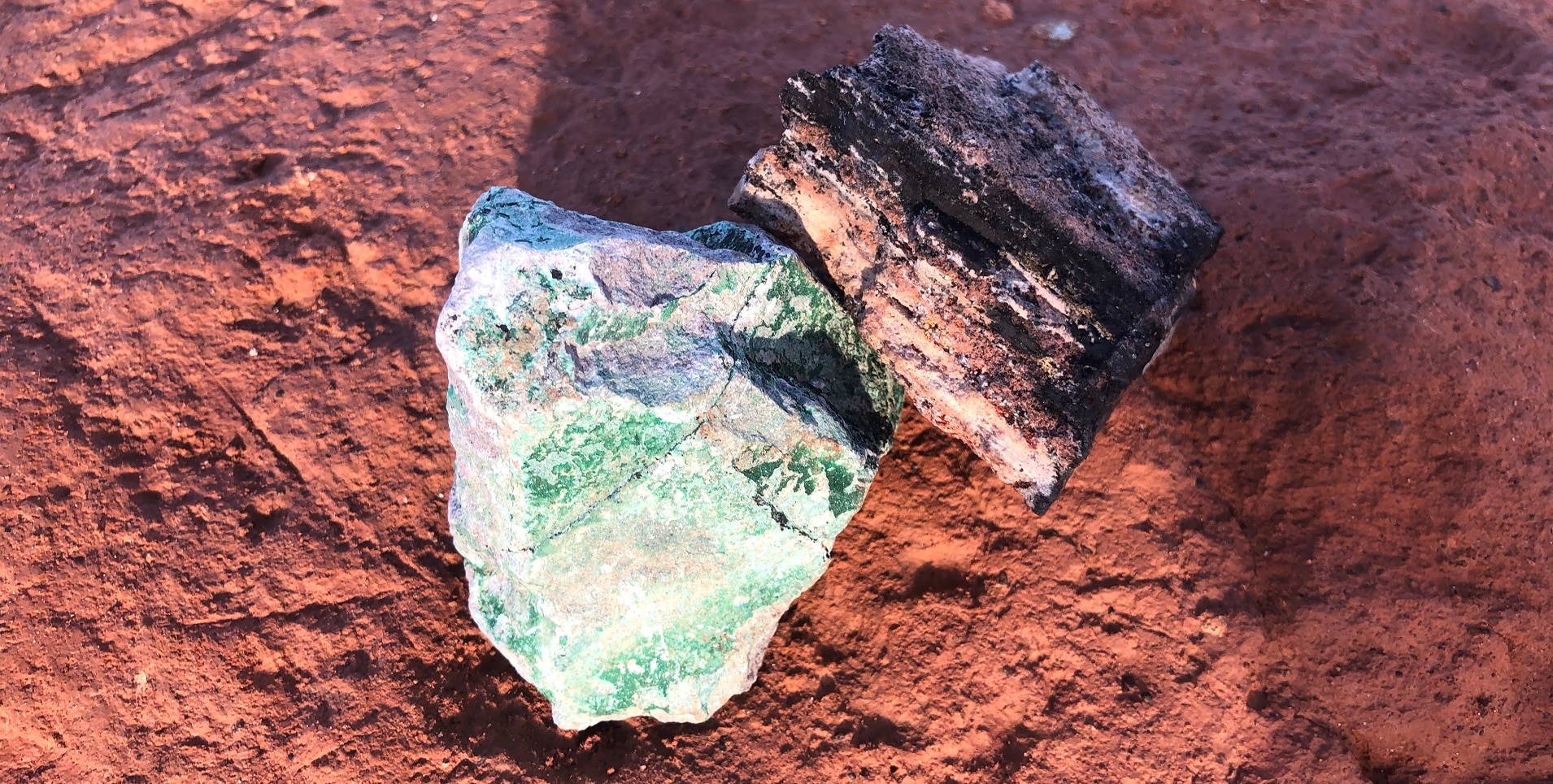 Un morceau de cobalt, de couleur noire, et un morceau de cuivre, de couleur verte, sont posés sur le sol ocre pour illustrer l’extraction minière dans la « Copper Belt » en République démocratique du Congo.