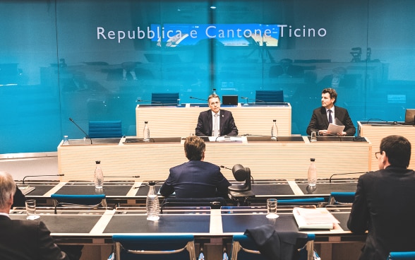 Le conseiller fédéral Cassis est assis en face des membres du Conseil d'État du Tessin. À sa droite, le secrétaire d'État Roberto Balzaretti. Sur le mur se trouve «Repubblica e Cantone Ticino».