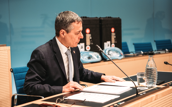 Le conseiller fédéral Cassis est assis à un bureau, les dossiers sont sur la table devant lui. En arrière-plan, vous pouvez voir les deux valises avec les respirateurs.