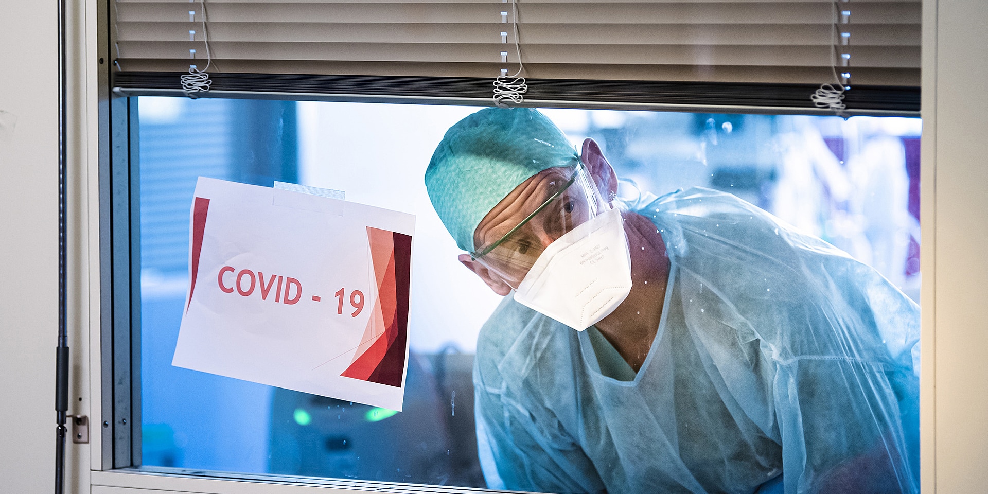   A health worker looks through an intensive care unit window past a taped-on paper sign that says 'COVID–19'. 