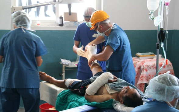 Drei Ärzte in blauen Kitteln kümmern sich um einen nepalesischen Patienten, der sich den Arm und das Bein gebrochen hat.