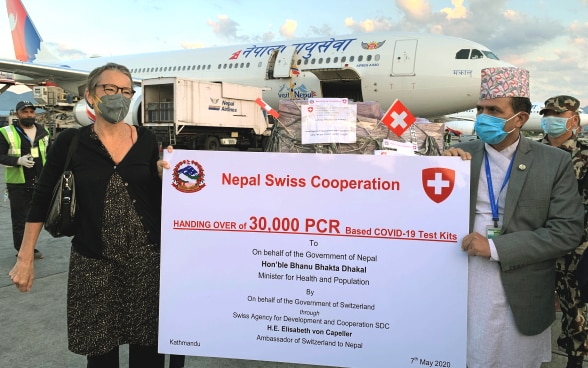 Die Schweizer Botschafterin in Nepal und Nepals Minister für Gesundheit und Bevölkerung stehen vor einem Flugzeug und halten ein Plakat in der Hand, in der die Übergabe von 30'000 COVID-19-Testkits angezeigt wird.