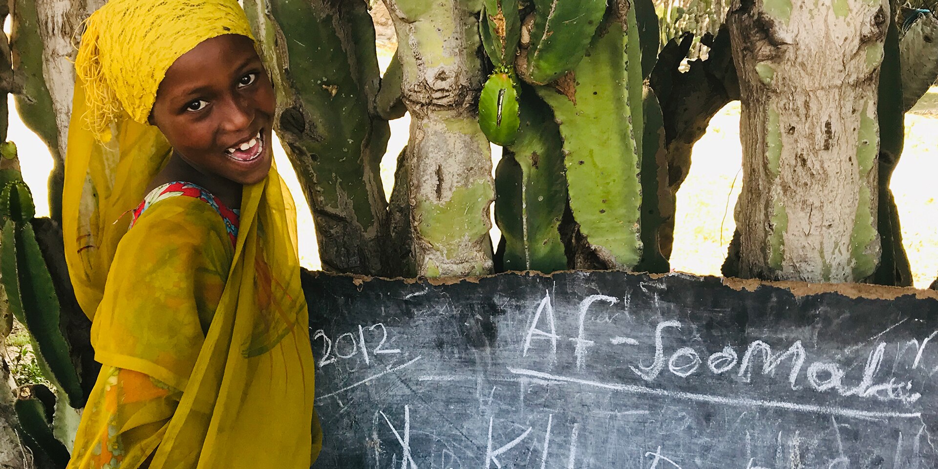  A little girl with a textbook sits at a wooden desk in a classroom and looks at the camera. 