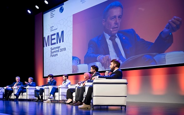Federal Councillor Ignazio Cassis speaking on a stage with young people sitting next to him at the MEM Forum in 2019. 
