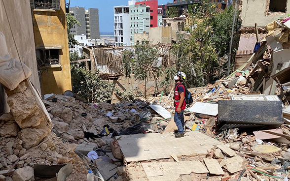 An expert from the Swiss Humanitarian Aid Unit is standing in front of a destroyed house surrounded by debris.