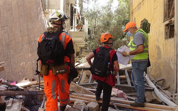 Three experts from the Swiss Humanitarian Aid Unit are standing in front of a destroyed house surrounded by debris.