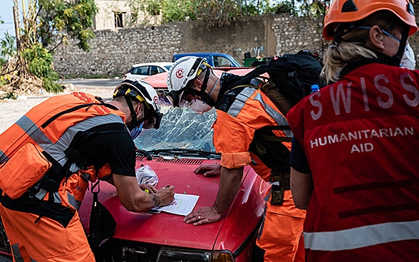 Drei Experten der Humanitären Hilfe des Bundes diskutieren einen Aktionsplan auf der Haube eines kaputten Fahrzeugs. 
