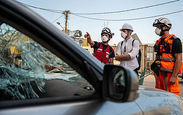 Blick durch ein zerbrochenes Fahrzeugfenster auf drei Experten der Humanitären Hilfe des Bundes. 