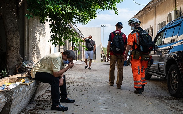 Un homme est assis sur le bord de la route, la tête dans les mains. Des experts du Corps suisse d'aide humanitaire passent à côté de lui.