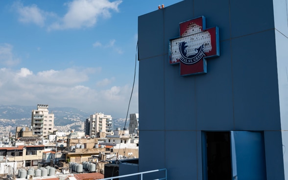 Blick auf das Äussere eines kaputten Spitals. Im Hintergrund sieht man die Folgen der Explosion auf die Stadt Beirut.