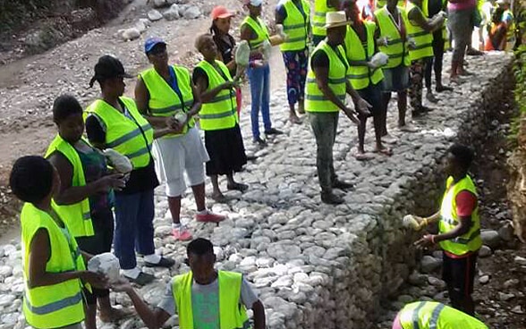 Plusieurs personnes portant un gilet de sécurité jaune déplacent des pierres pour construire des gabions.