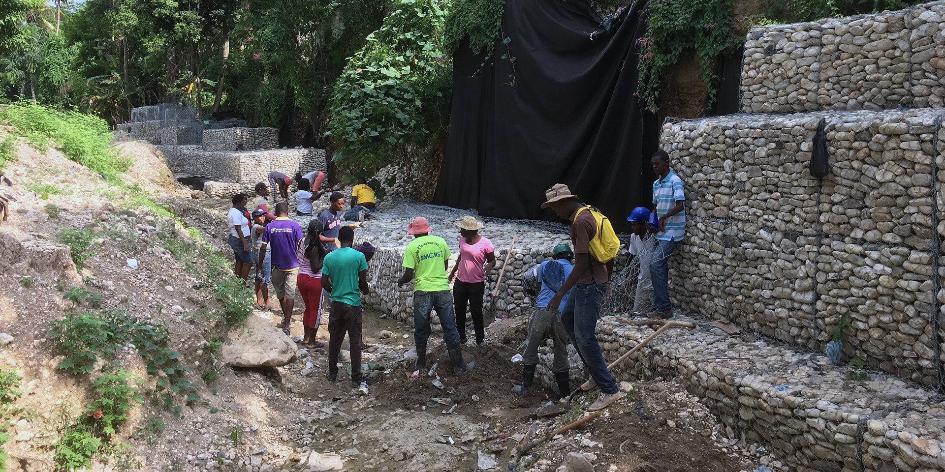 Un gruppo di persone lavora alla costruzione di strutture in pietra lungo una via, che formano alti argini per incanalare l’acqua.