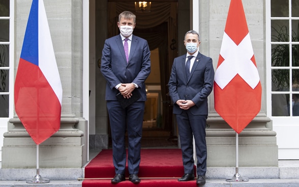 Ignazio Cassis et Tomáš Petříček posent debout, à l’entrée d’un bâtiment. À côté d’eux, les drapeaux de leurs pays respectifs.