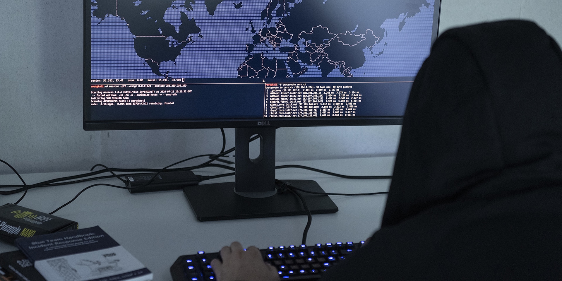 Photo symbolisant la cybercriminalité: assis devant un écran d’ordinateur montrant une carte du monde, un homme dont la tête est couverte d’une capuche modifie des codes de programmation.
