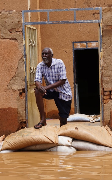 A man is standing on some sacks placed in front of his house half destroyed. The house overlooks the road submerged by water.