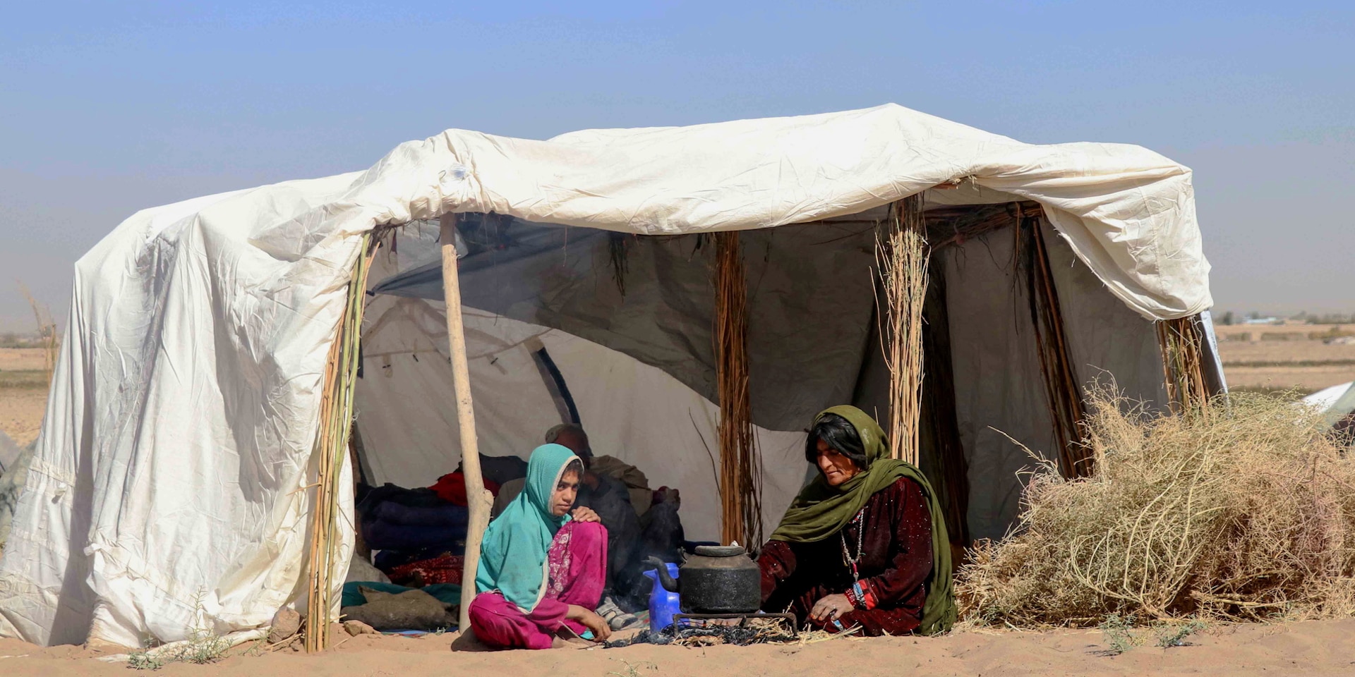 A woman and a young girl sit under a makeshift tent in the desert.