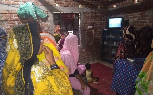 A group of young girls sit together in a hall and watch a televised puppet show.