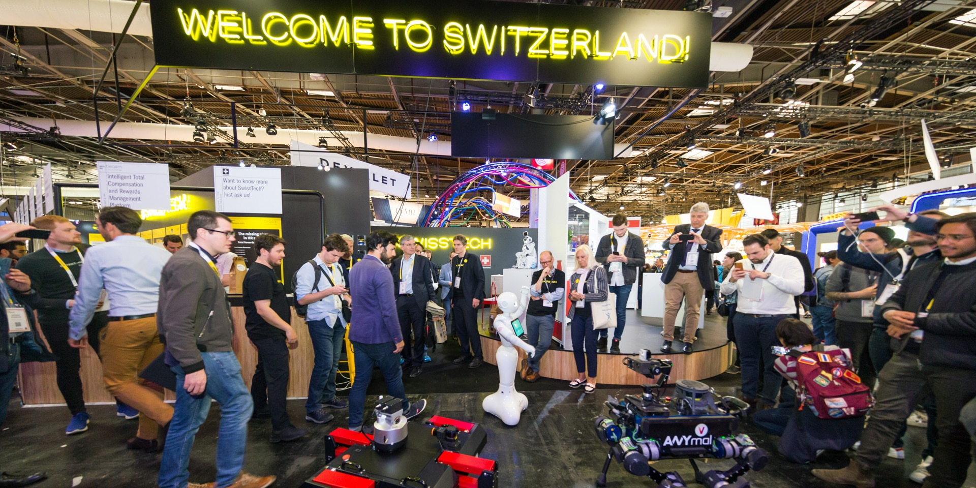 Dans une salle d'exposition, les gens regardent des drones, des robots et des technologies innovantes. Au-dessus, se trouve un panneau sur lequel est inscrit «Bienvenue en Suisse».
