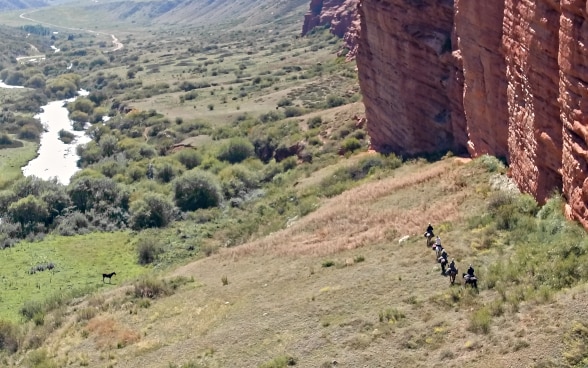 Five riders trek along a rock face in a remote valley.