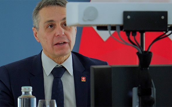 Le conseiller fédéral Ignazio Cassis prononce un discours face à une caméra lors d’une conférence virtuelle. Derrière lui, le drapeau de la Suisse.