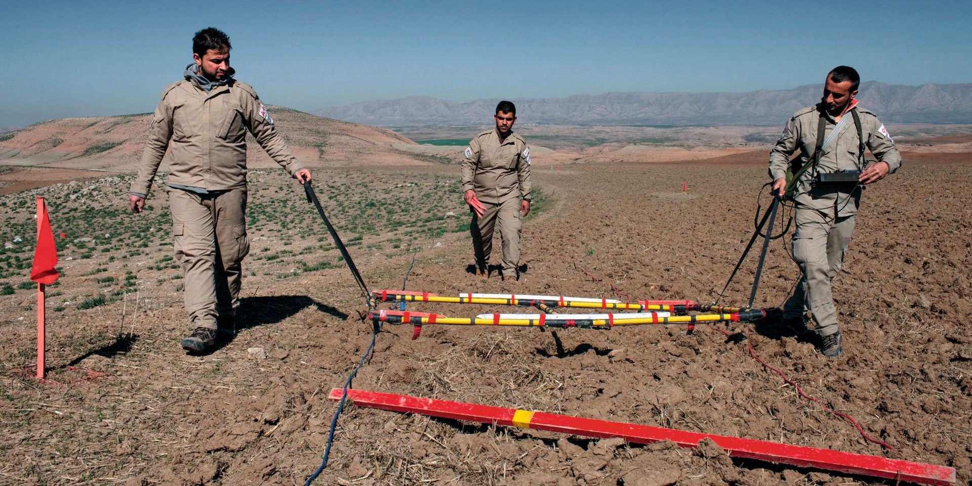  Trois hommes en combinaison procèdent au déminage d’un terrain dans le désert iraqien. 