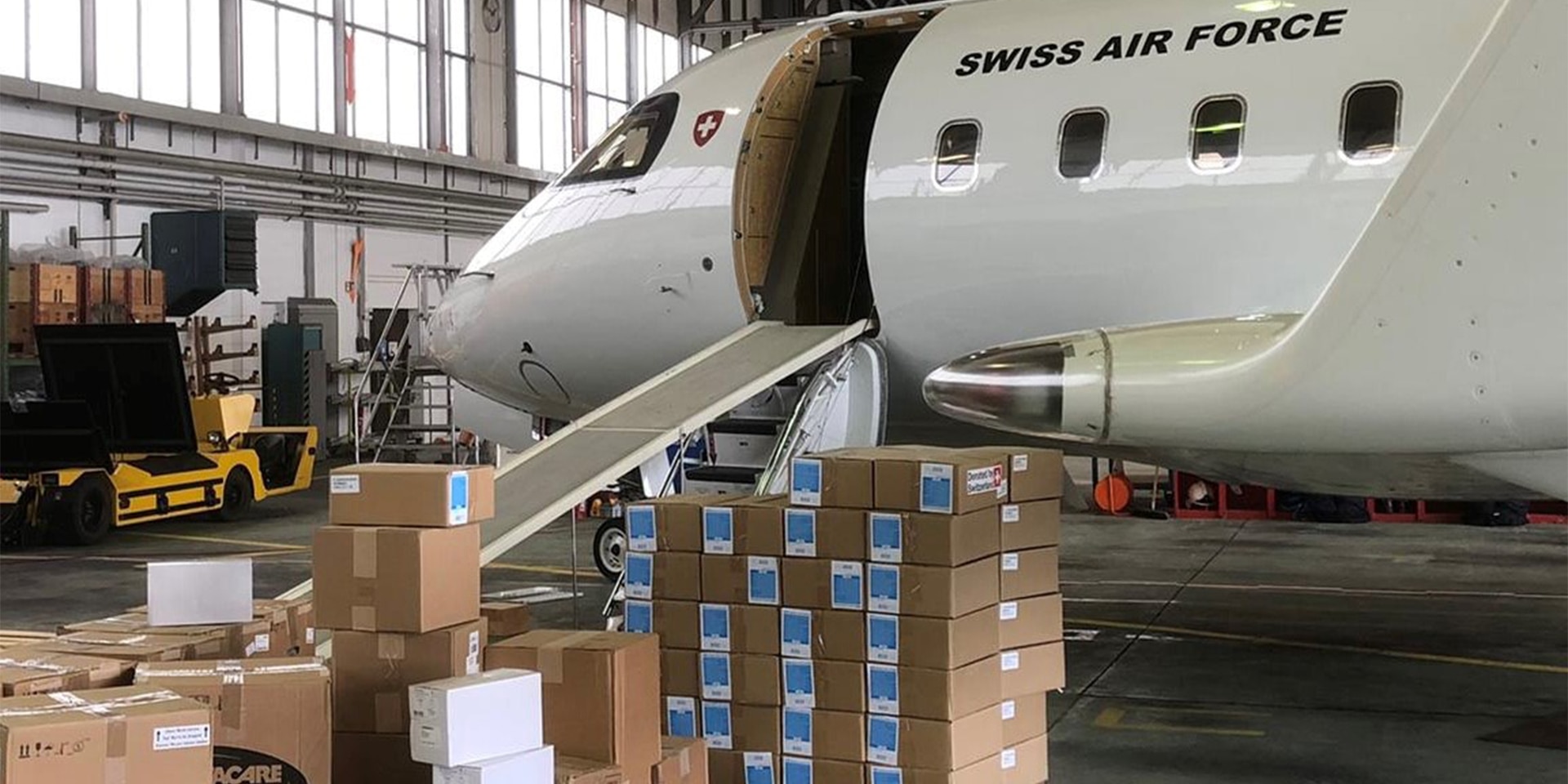 Im Bild sieht man ein Flugzeug der Schweizer Eidgenossenschaft im Hangar stehen. Davor stehen Kisten mit Hilfsgütern.