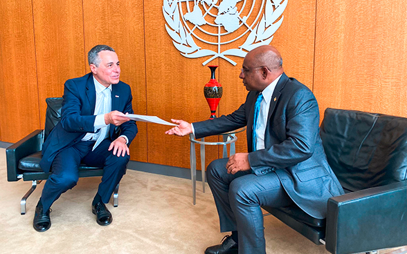 Avec l'emblème de l'ONU en toile de fond, le conseiller fédéral Ignazio Cassis remet un document au président de l'Assemblée générale de l'ONU Abdulla Shahid (tous deux assis).