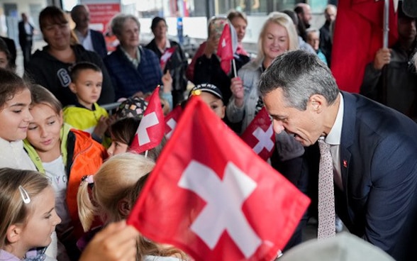 Le quai de la gare d'Airolo est bondé pour accueillir le train spécial. Le président de la Confédération Ignazio Cassis salue les personnes présentes pendant que des écoliers agitent des drapeaux suisses.