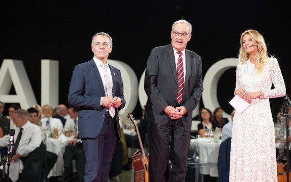 Ignazio Cassis, Guy Parmelin e Christa Rigozzi parlano su un palco.