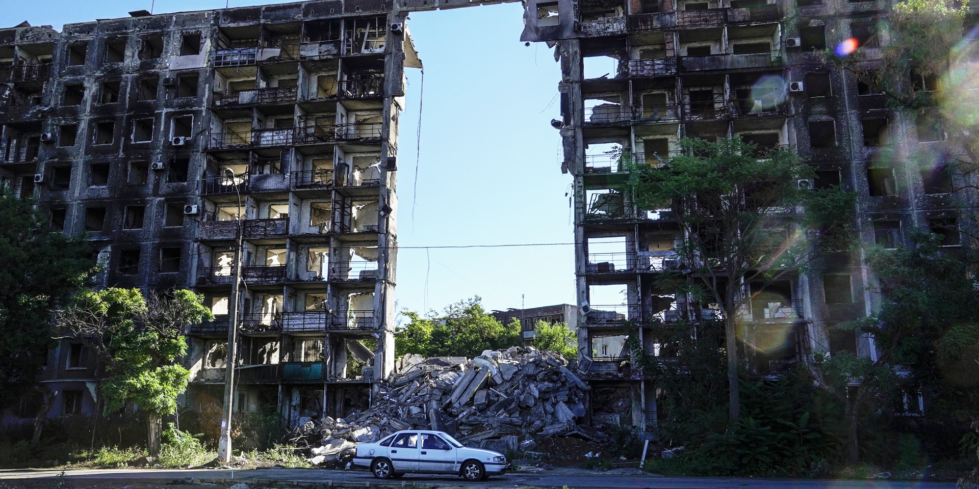 A white car in front of housing blocks destroyed by shelling. A blue sky is visible through a large gap in the blocks.